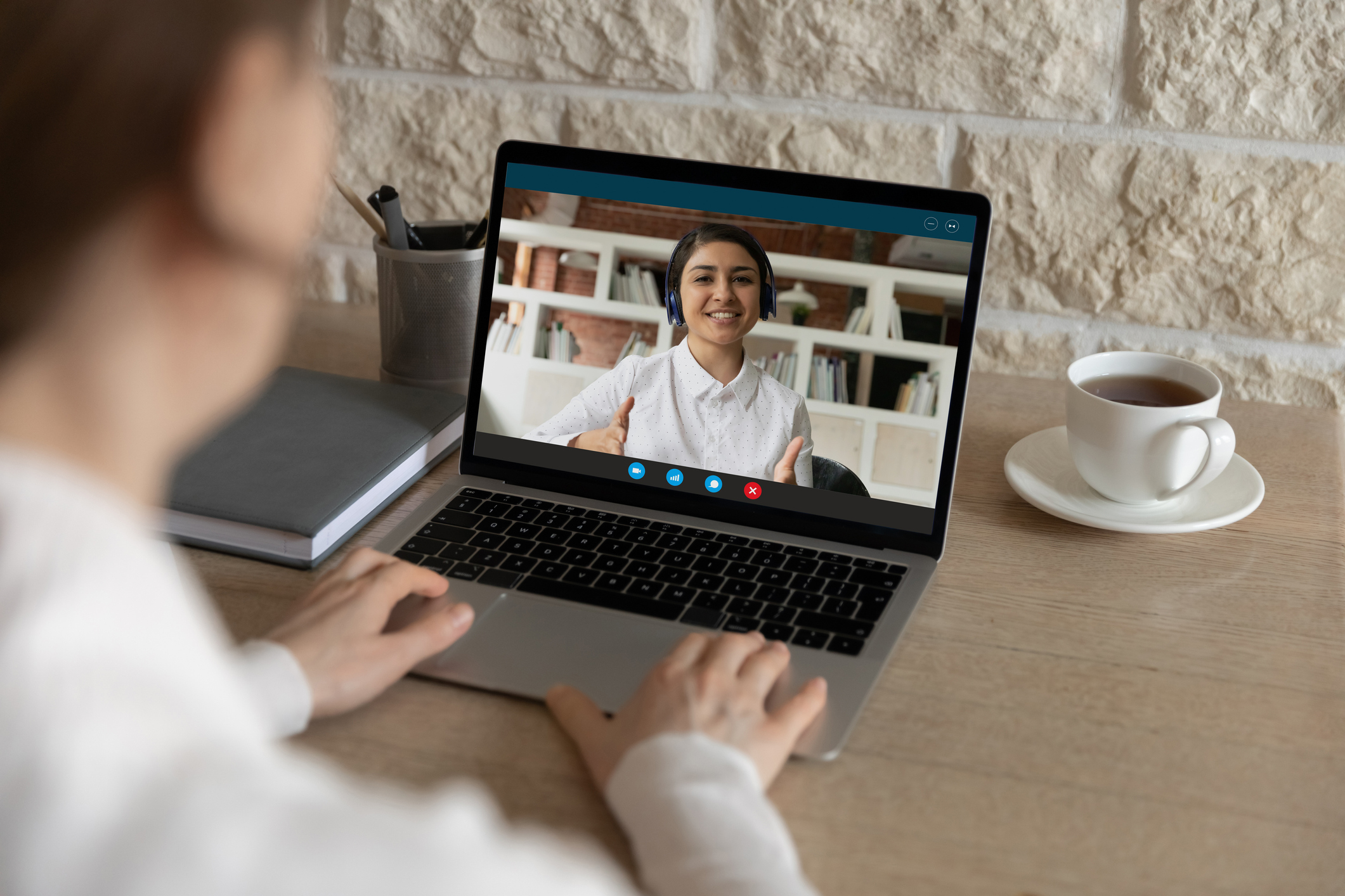 A person sits at a wooden desk using a laptop for a video call, with the screen showing another person waving from what appears to be a home office with bookshelves in the background. A coffee cup and pen holder are visible on the desk beside the laptop.
