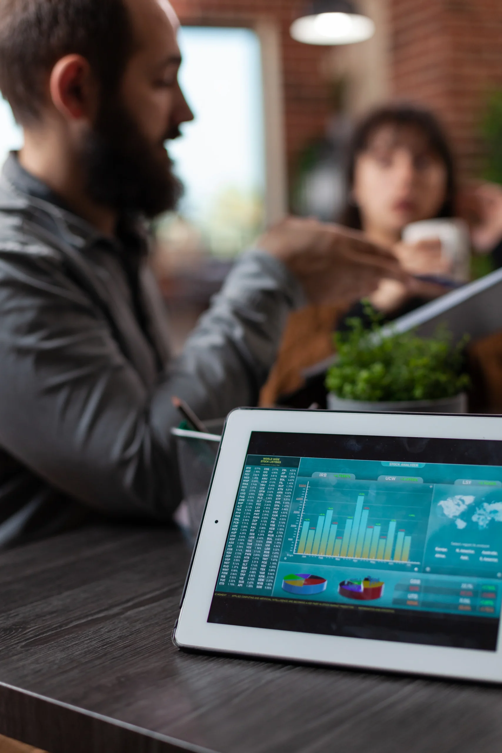 A bearded man in a gray shirt sits at a wooden table working on a tablet displaying colorful business analytics charts and data visualizations, with another person blurred in the background in what appears to be a brick-walled office or cafe setting.