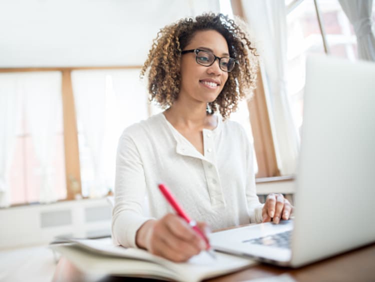 A smiling woman with curly hair and glasses sits at a desk writing in a notebook while working on a laptop computer in a bright, modern office space.