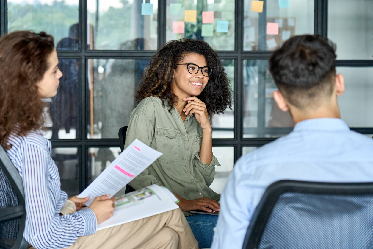 Three professionals collaborate in a modern office setting, with a woman in glasses and olive green shirt smiling while discussing documents with two colleagues seated around a table near large windows with colorful sticky notes.