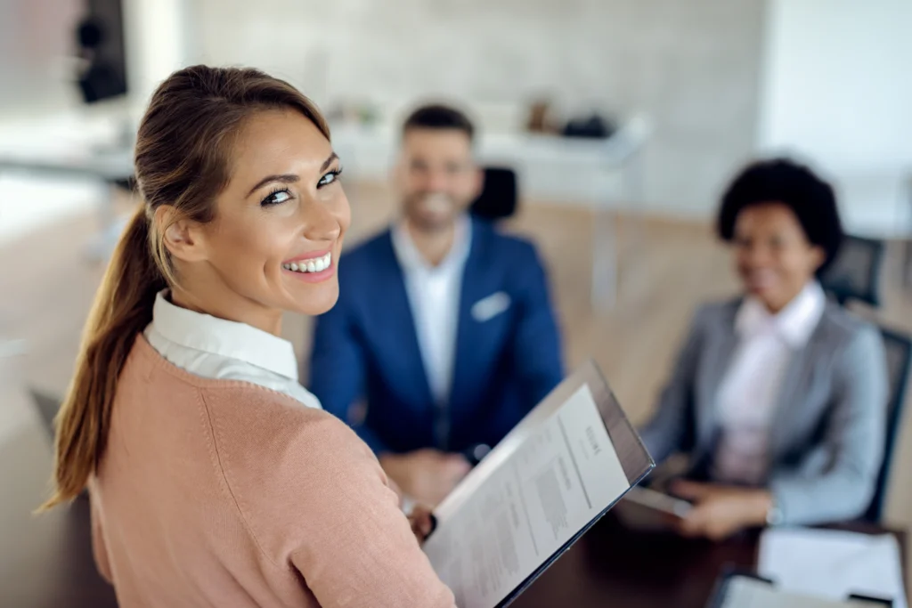 A smiling woman in a beige sweater holds documents while looking over her shoulder during a business meeting, with two colleagues visible in the blurred background of a modern office setting.