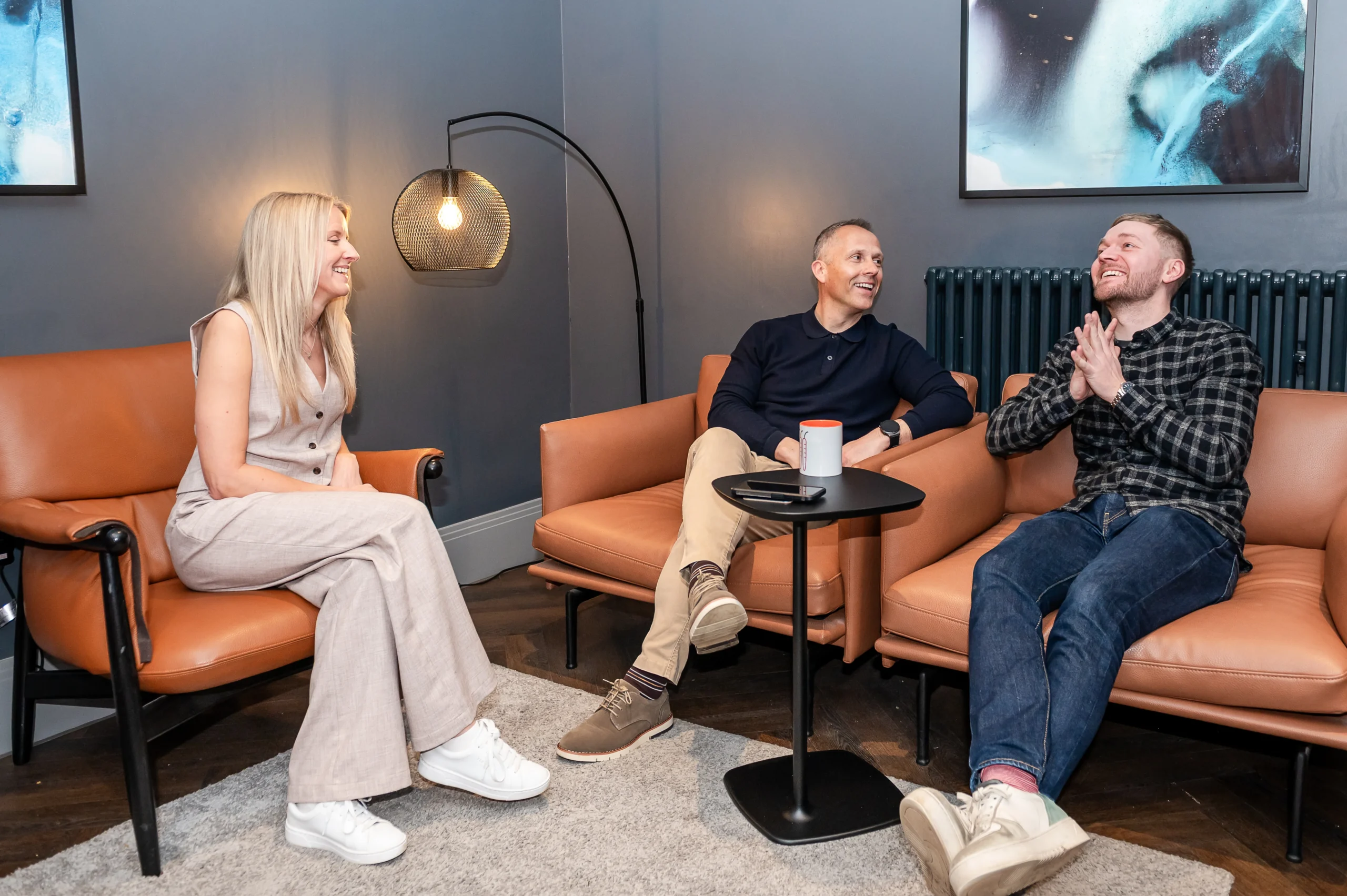 Three people sit on orange leather chairs in a modern lounge setting, engaged in conversation. A blonde woman in white sits across from two men, with a small black table and floor lamp between them, and abstract blue artwork visible on the gray walls behind them.